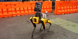 a yellow robot dog, called spot, stands in the middle of a room in an industrial setting it has an ipad on its "head" to show a person talking