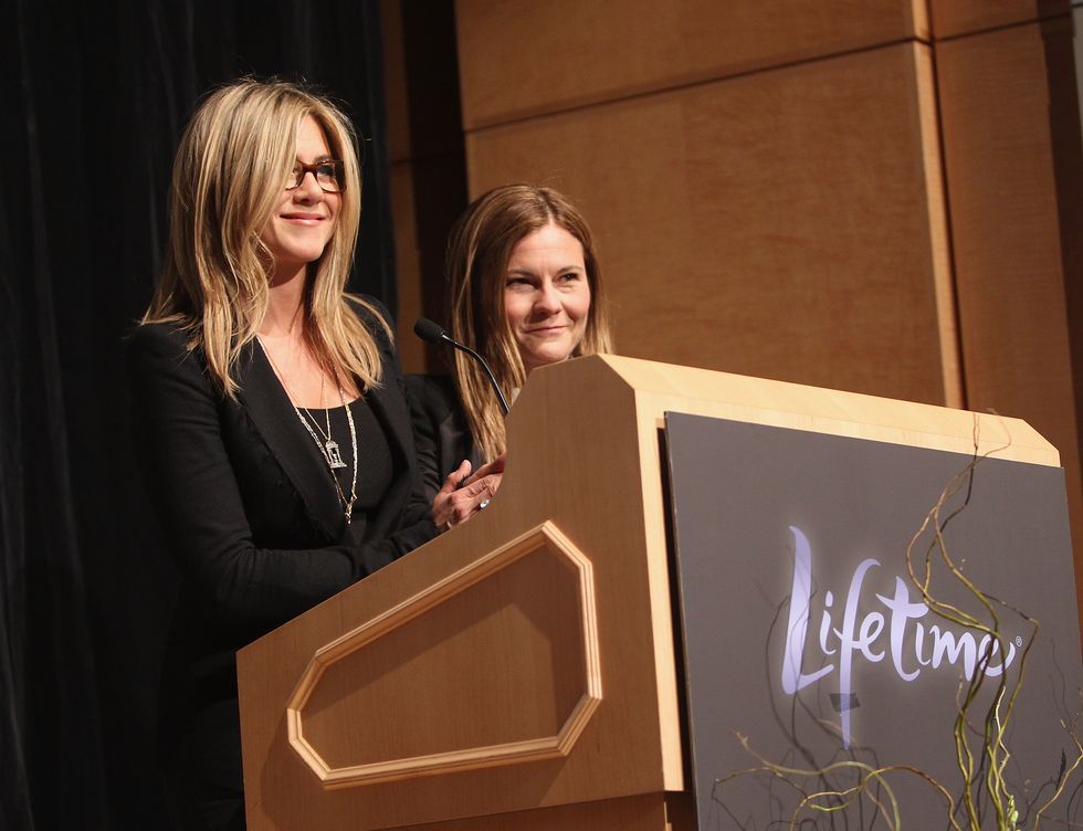 jennifer aniston and kristin hahn stand behind a light wooden podium with a lifetime logo on the front, both smile as they look out toward an audience