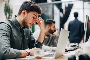 exhausted businessman looking at laptop on desk while sitting with male coworker in office