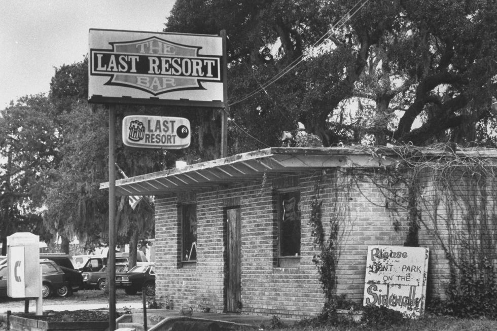 a brick building has a large sign outside advertising the last resort bar, several large trees are in the background as well as a pay phone and parked cars