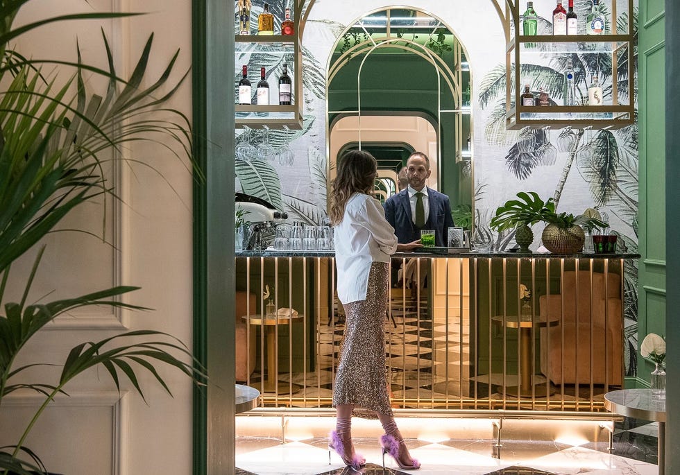 woman in skirt and high heels standing next to a bar counter at a hotel, with gilded finishes, palm tree wallpaper in back, and a man in a tux working