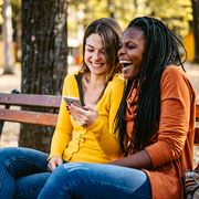 fall autumn jokes two friends sitting on a park bench looking at a phone and laughing together