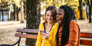 fall autumn jokes two friends sitting on a park bench looking at a phone and laughing together