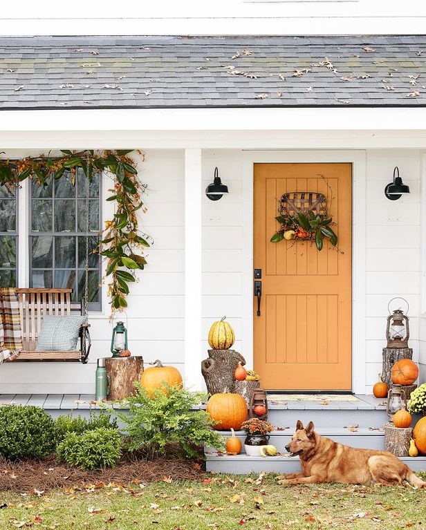 a white cottage with an orange door with a wreath and stacks of pumpkins and mums