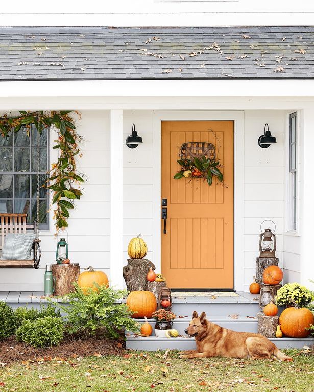 a white cottage with an orange door with a wreath and stacks of pumpkins and mums