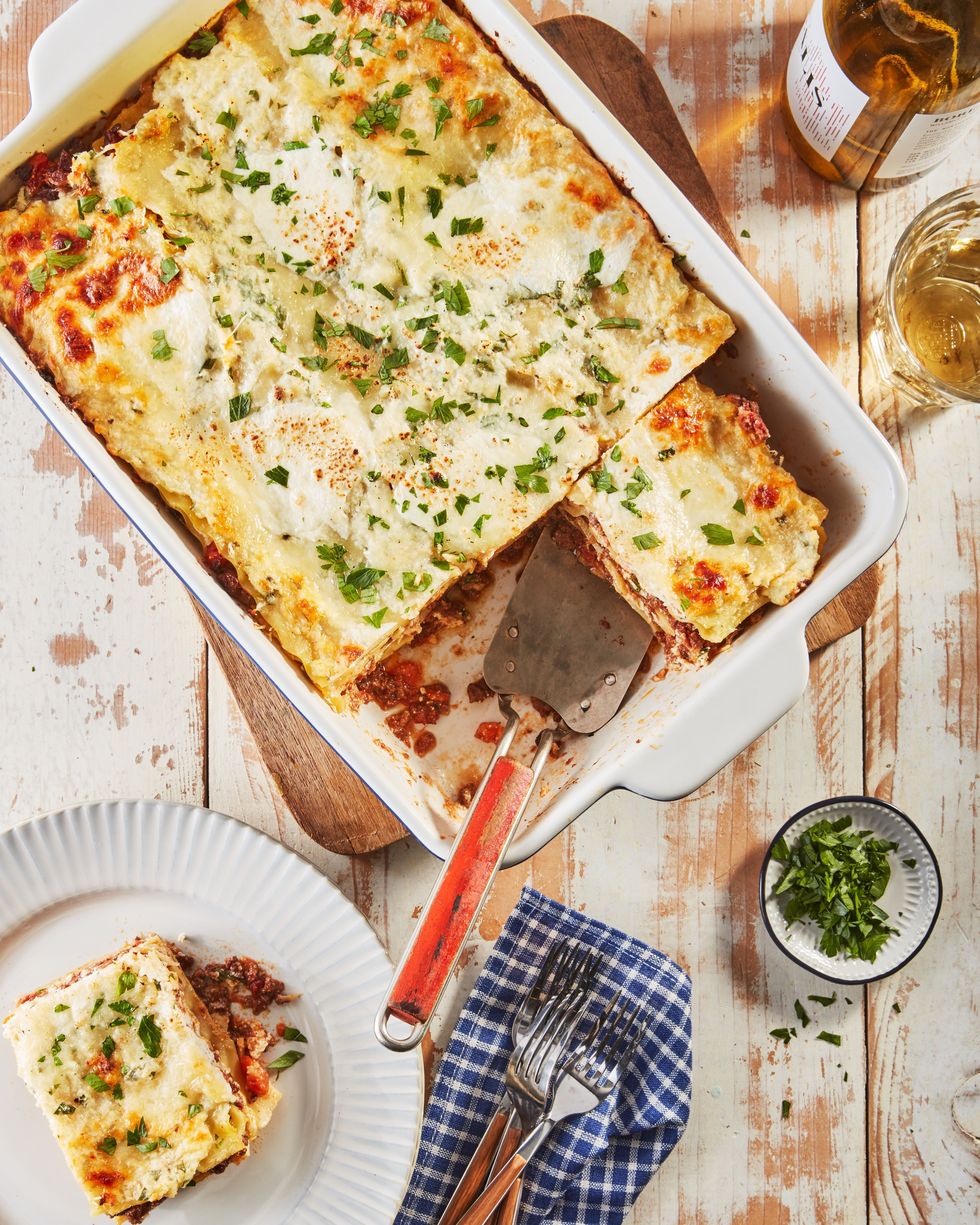lasagna bolognese in a white square baking dish with a red spatula lifting a portion out and another portion on a white plate next to it