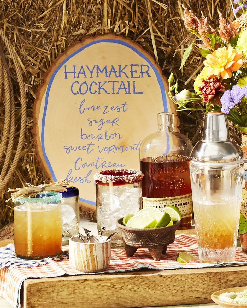 a wooden table outside with one cocktail and a cup of ice and a small bowl of lime wedges and a cocktail shaker and a bottle of bourbon