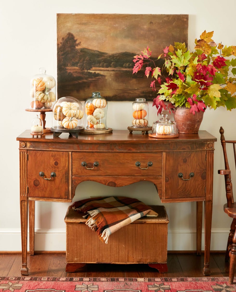 bouquet of colorful fall branches in rustic clay vessel, atop sideboard beside mini pumpkins displayed under glass cloches