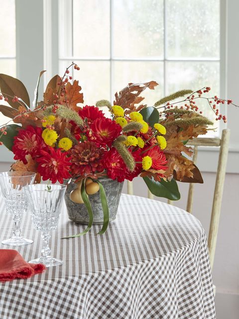 red and yellow floral arrangement in galvanized pale embellished with dried fall leaves, displayed on gray gingham tablecloth
