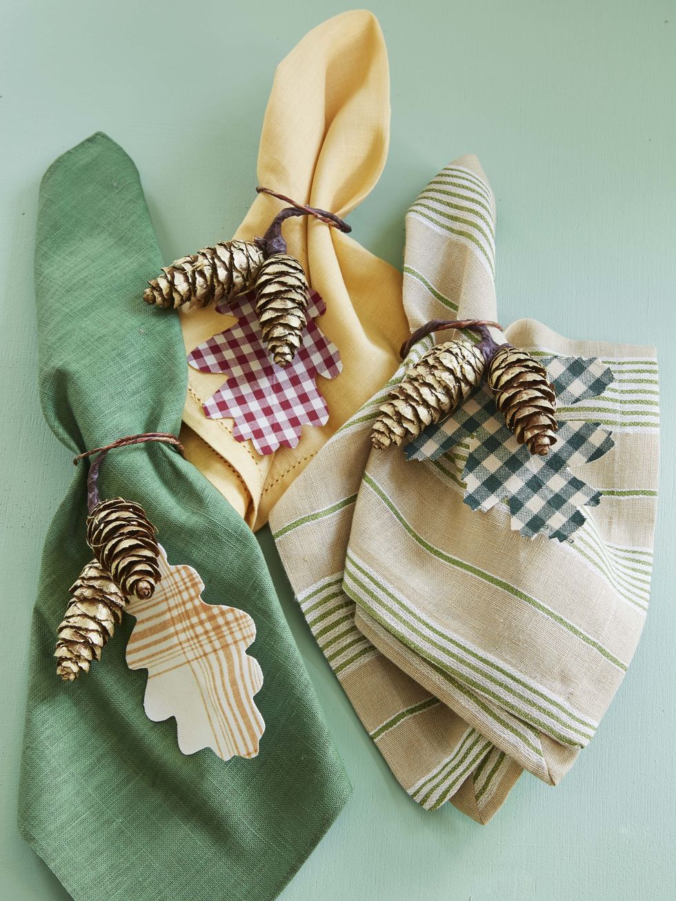 three napkin rings crafted from twine, pinecones, and fall leaf shapes cut from gingham and plaid fabrics