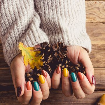 closeup of a young womans hands with trendy fall nail colors yellow, turquoise, and brown she wears a cozy gray knit sweater, resting her hands on a rustic table adorned with leaves, pinecones, acorns