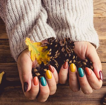 closeup of a young womans hands with trendy fall nail colors yellow, turquoise, and brown she wears a cozy gray knit sweater, resting her hands on a rustic table adorned with leaves, pinecones, acorns