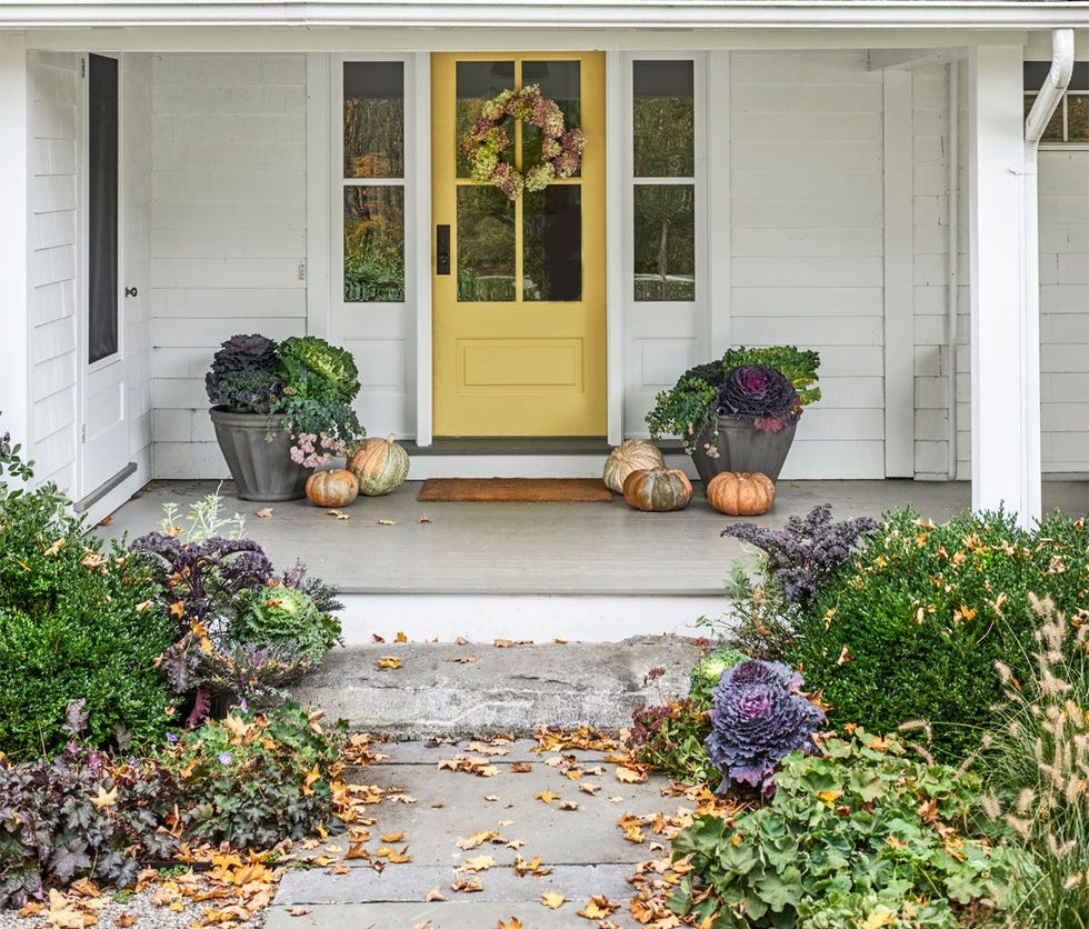 white farmhouse with covered porch and yellow front door decorated for fall with dried hydrangea wreath, pumpkins cabbages