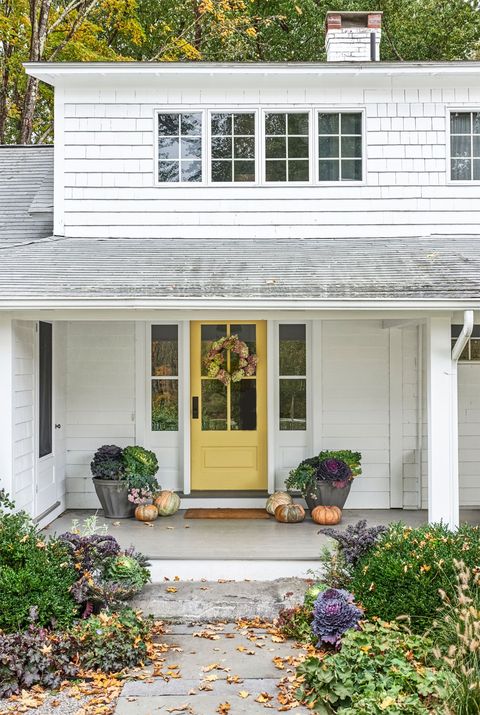 white farmhouse with covered porch and yellow front door decorated for fall with dried hydrangea wreath, pumpkins cabbages
