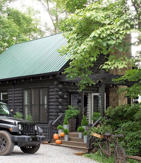 pumpkins and green plants in metal planters decorate front porch steps of log cabin with green roof and jeep parked out front