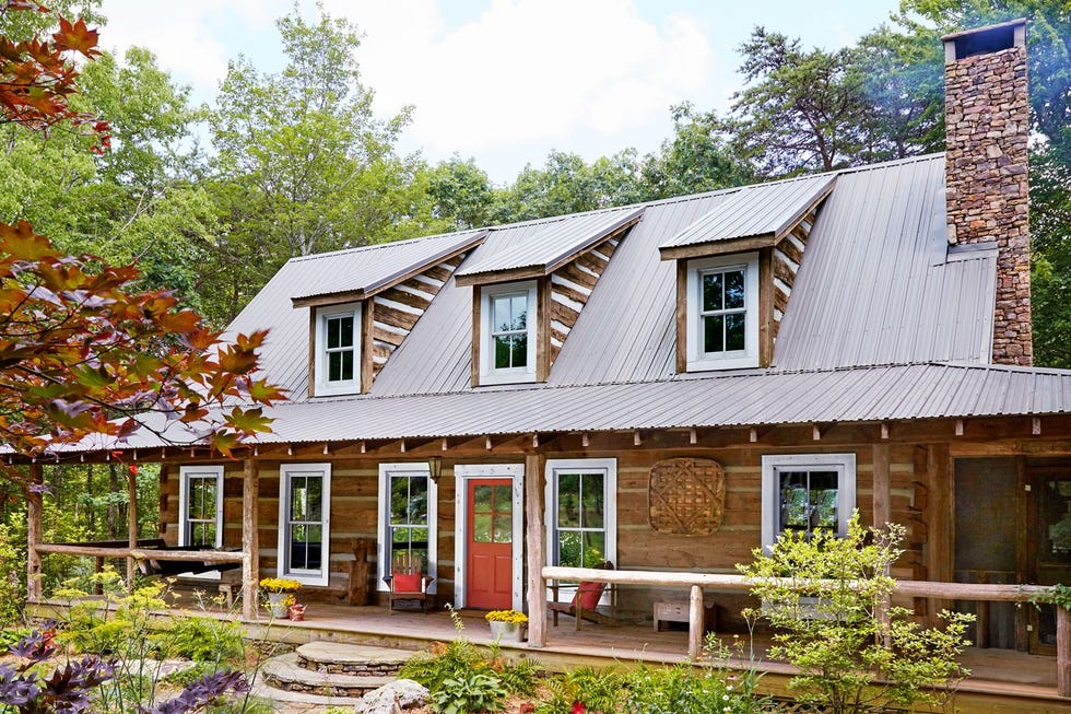 log cabin with a long front porch and an orange front door decorated for fall with yellow mums in galvanized buckets