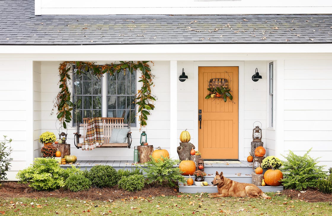 white farmhouse fall porch decor with orange door, plaid throw blanket on swing, magnolia garland, tobacco basket door wreath