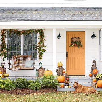 white farmhouse fall porch decor with orange door, plaid throw blanket on swing, magnolia garland, tobacco basket door wreath