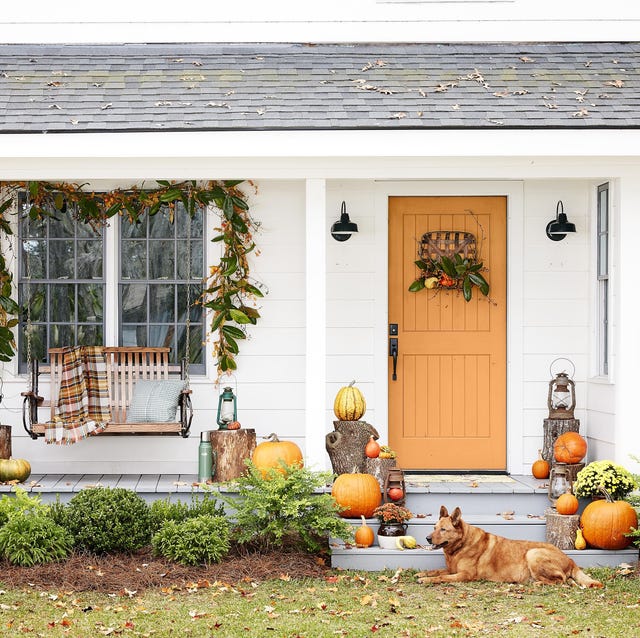 white farmhouse fall porch decor with orange door, plaid throw blanket on swing, magnolia garland, tobacco basket door wreath