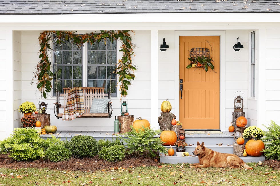 white farmhouse fall porch decor with orange door, plaid throw blanket on swing, magnolia garland, tobacco basket door wreath