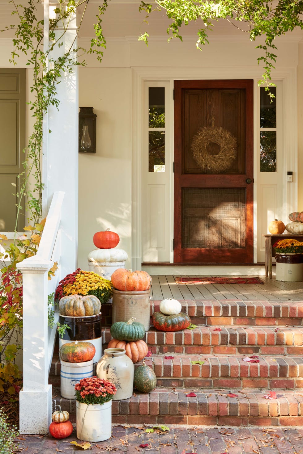 pumpkins and mums in vintage crocks arranged on brick steps along chunky white wood railing leading to a covered front porch