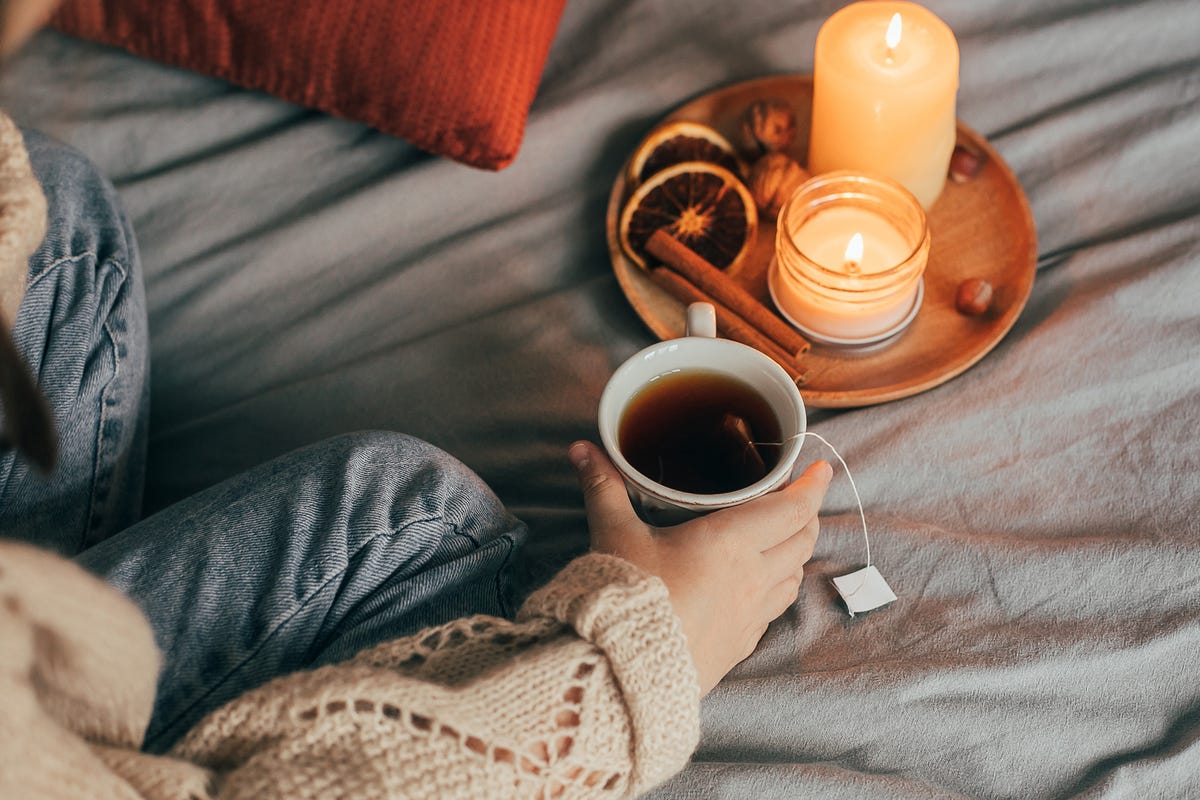 person holding tea in bed with candles, cinnamon sticks and dried oranges