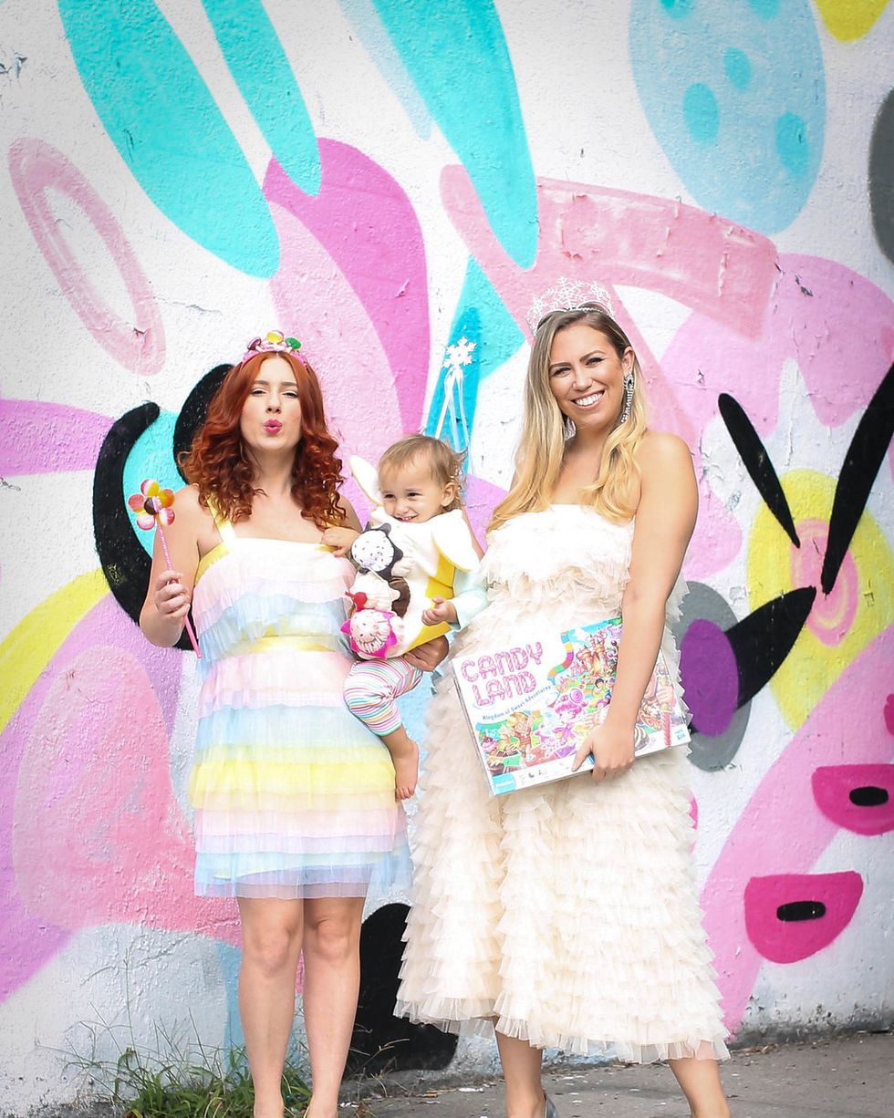 two friends dressed as candy land characters and baby in banana split costume standing in front of colorful mural