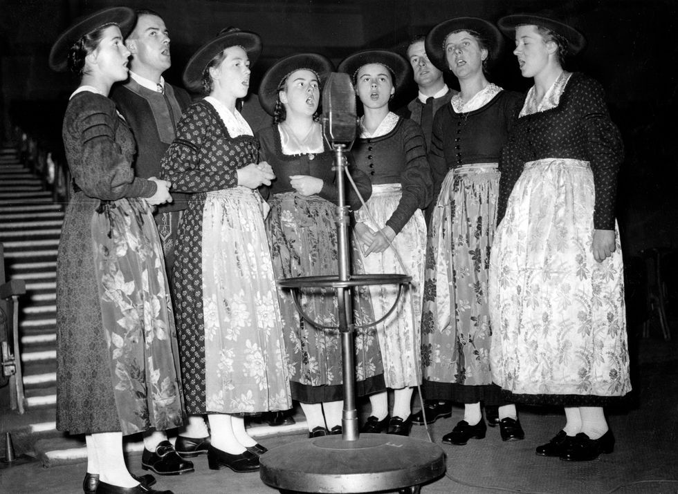 eight members of the von trapp family stand around a large microphone and sing, the girls wear dresses with matching aprons and hats, the men wear matching tops