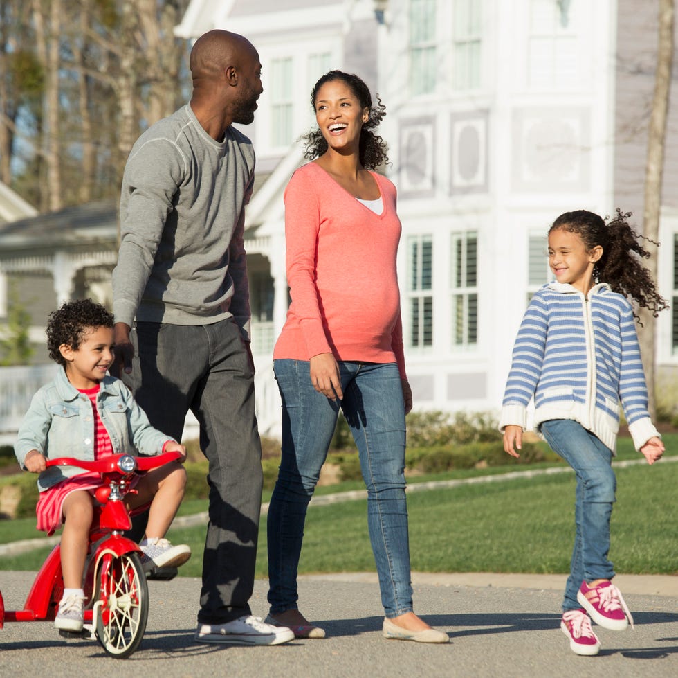 a black family with one child riding a red tricycle and a man and woman and little girl walking together on a sunny suburban street