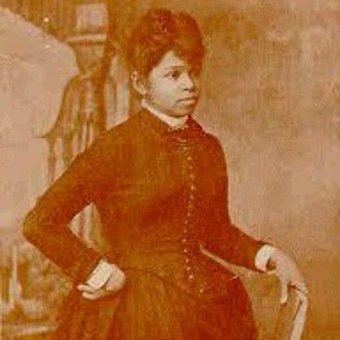 sarah boone stands for a portrait, she wears a dark dress with several buttons on the torso, her left hand rests on a chair back and her right hand rests on her hip with her elbow out