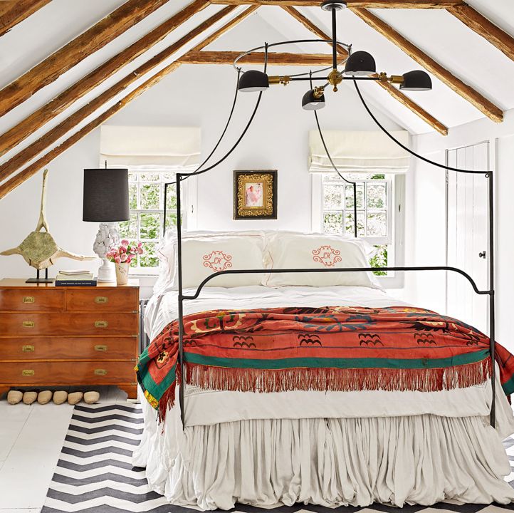 a wrought iron canopy bed complements the steep pitched ceiling in this upstairs bedroom with white walls and a wooden dresser and red blanket on the bed