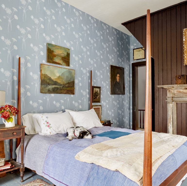 a farmhouse bedroom that has an statement accent wall in a light blue with white flowers wallpaper and theres a pencil post bed with a little black and white dog on it