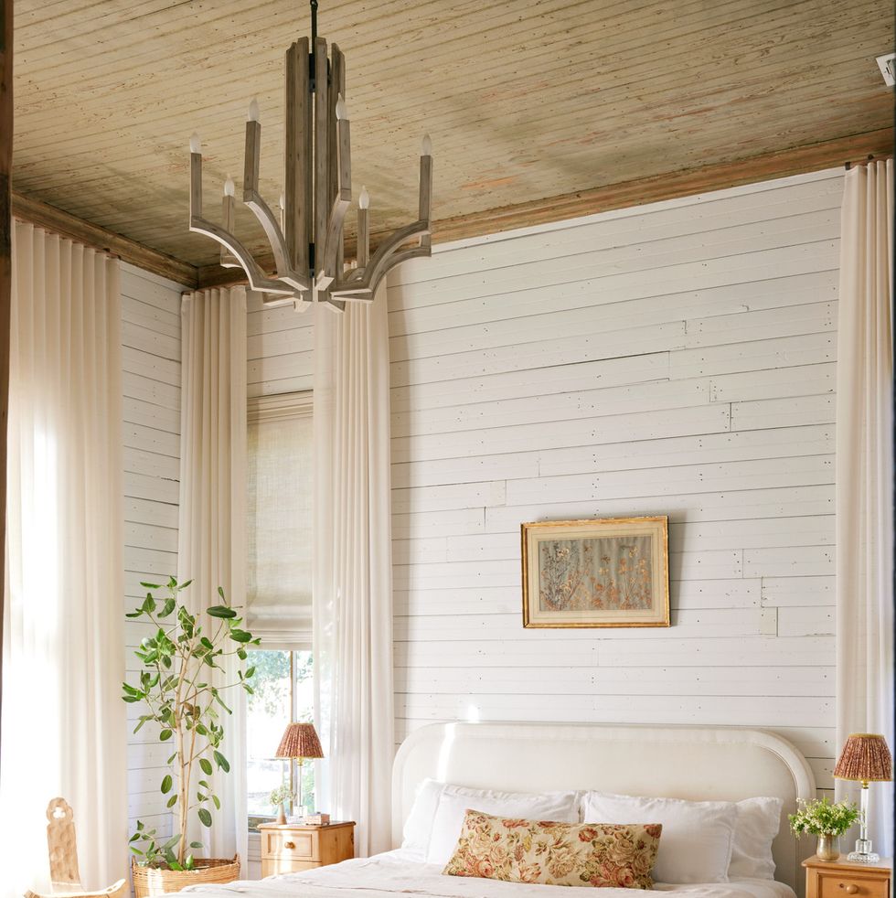 linen draperies hang in a bedroom against white shiplap walls and the bed is all white too with a floral pillow