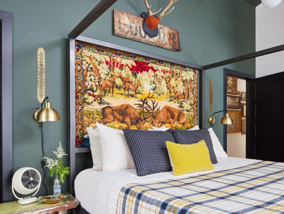 a vintage wildlife themed tapestry stands in as a headboard in a bedroom and the walls are a gray green color and the bed is made with plaid bedding and yellow and navy blue pillows