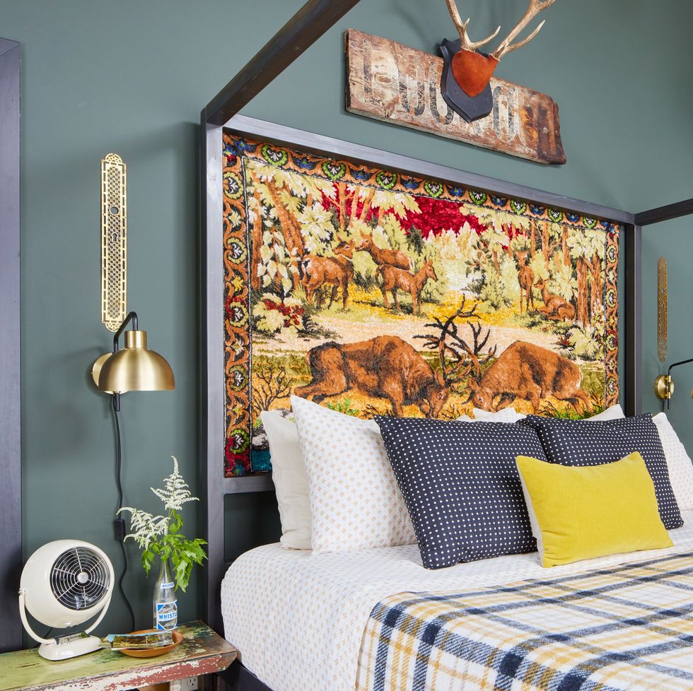 a vintage wildlife themed tapestry stands in as a headboard in a bedroom and the walls are a gray green color and the bed is made with plaid bedding and yellow and navy blue pillows