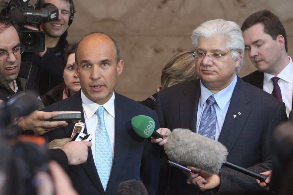 jim balsillie, wearing a black suit and blue tie, and mike lazaridis, wearing a dark blue suit and light blue shirt and tie, stand among several reporters, some of whom place microphones in front of them