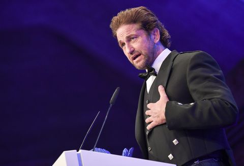 gerard butler wearing a black tuxedo, speaking into a microphone at a podium
