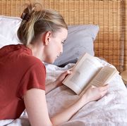 woman wearing a red shirt and jeans lying on bed reading a feel good book in sunlight
