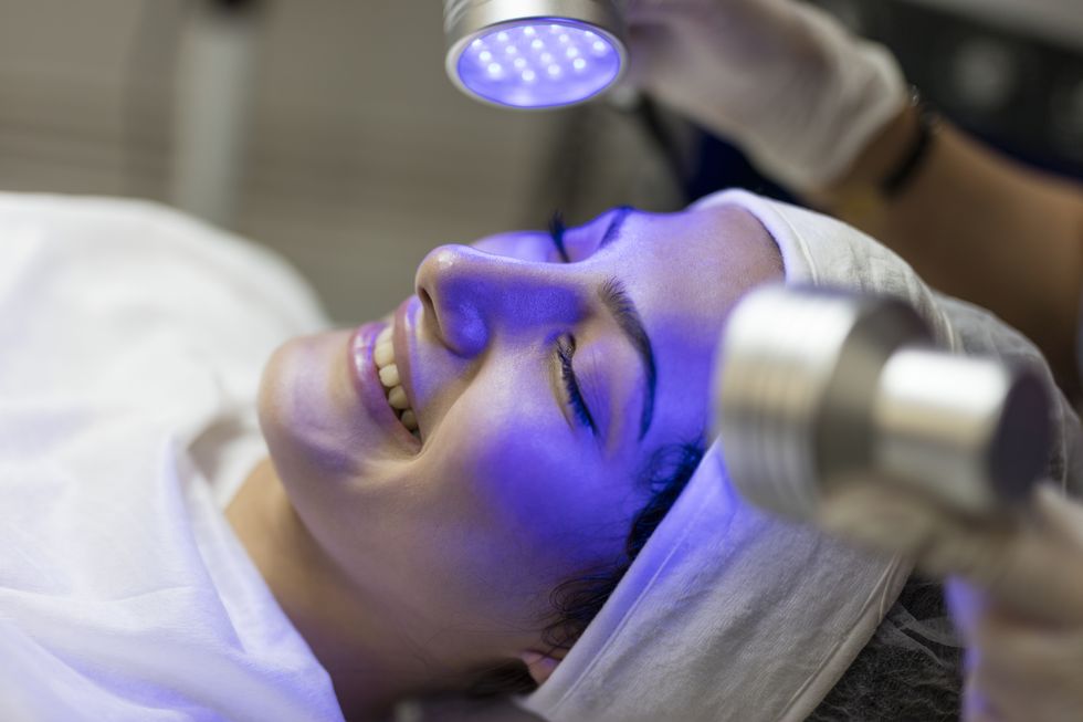 female beautician doing blue light therapy on face of pretty young woman