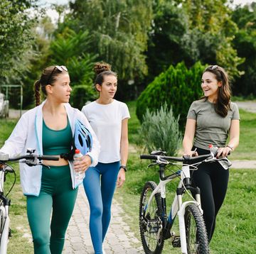 female friend group dressed in sporty clothes walking and talking while pushing bikes in city park