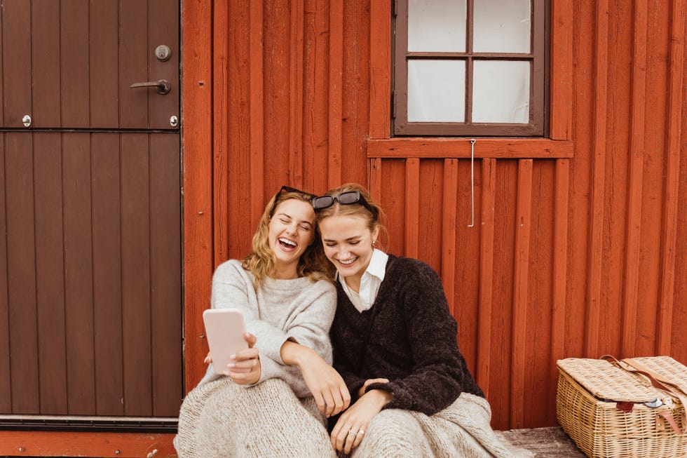 female friends laughing while taking selfie on mobile phone against cottage