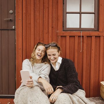 female friends laughing while taking selfie on mobile phone against cottage