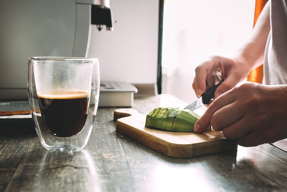 female hand cutting avocado on a wooden cutting board for toasts glass mug with hot coffee