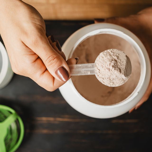 female hand holds measuring spoon with portion of whey protein powder above plastic jar on wooden table with shaker, banana and apple fruit process of making protein drink, top view