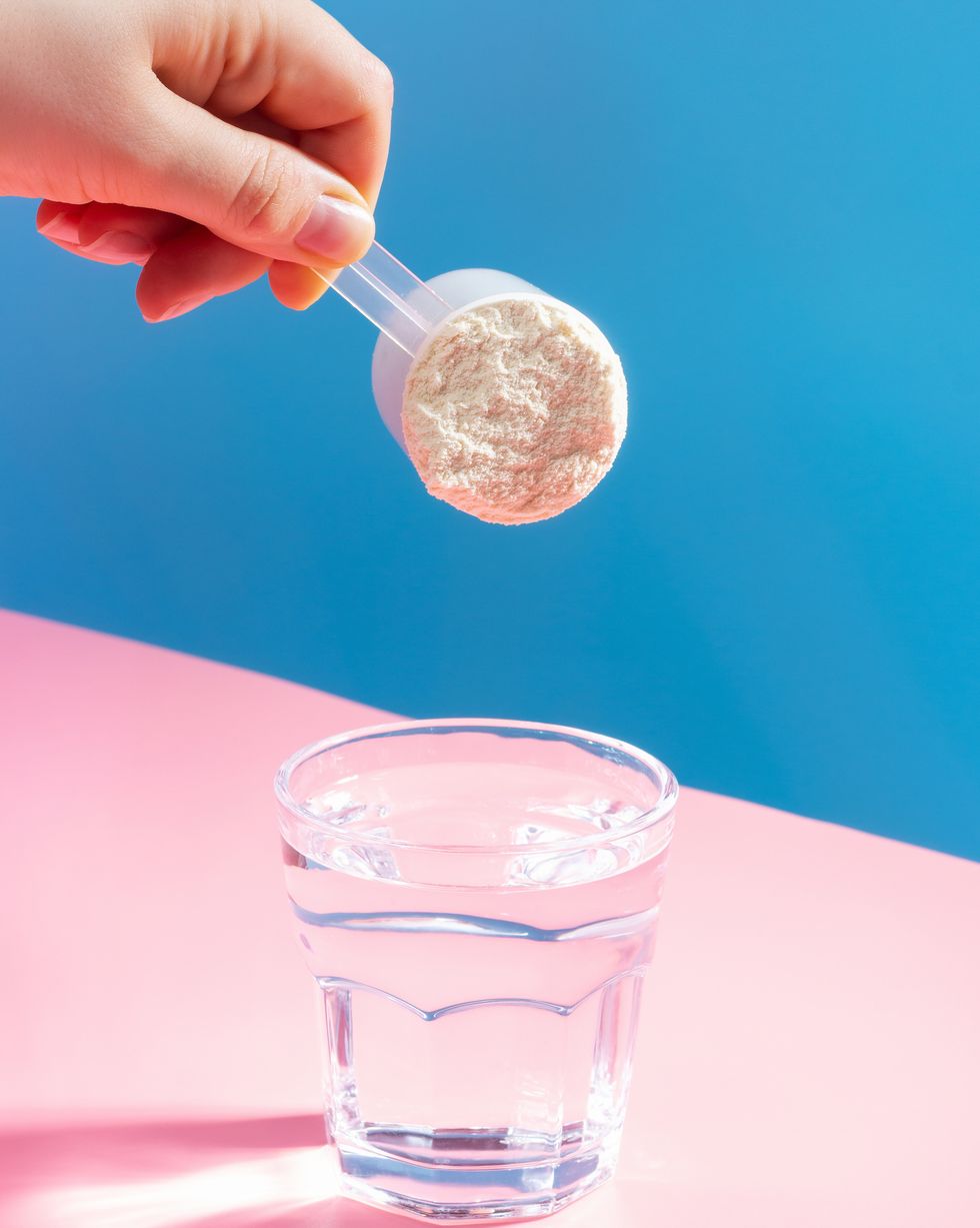 female hand pouring protein powder into glass of water on blue pink background