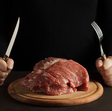 twee handen met mes en vork bij een grote homp rauw vlees op een snijplank