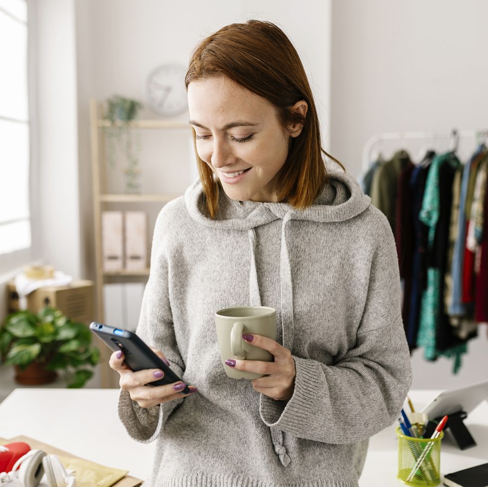 female owner using cell phone and drinking coffee while standing in home office