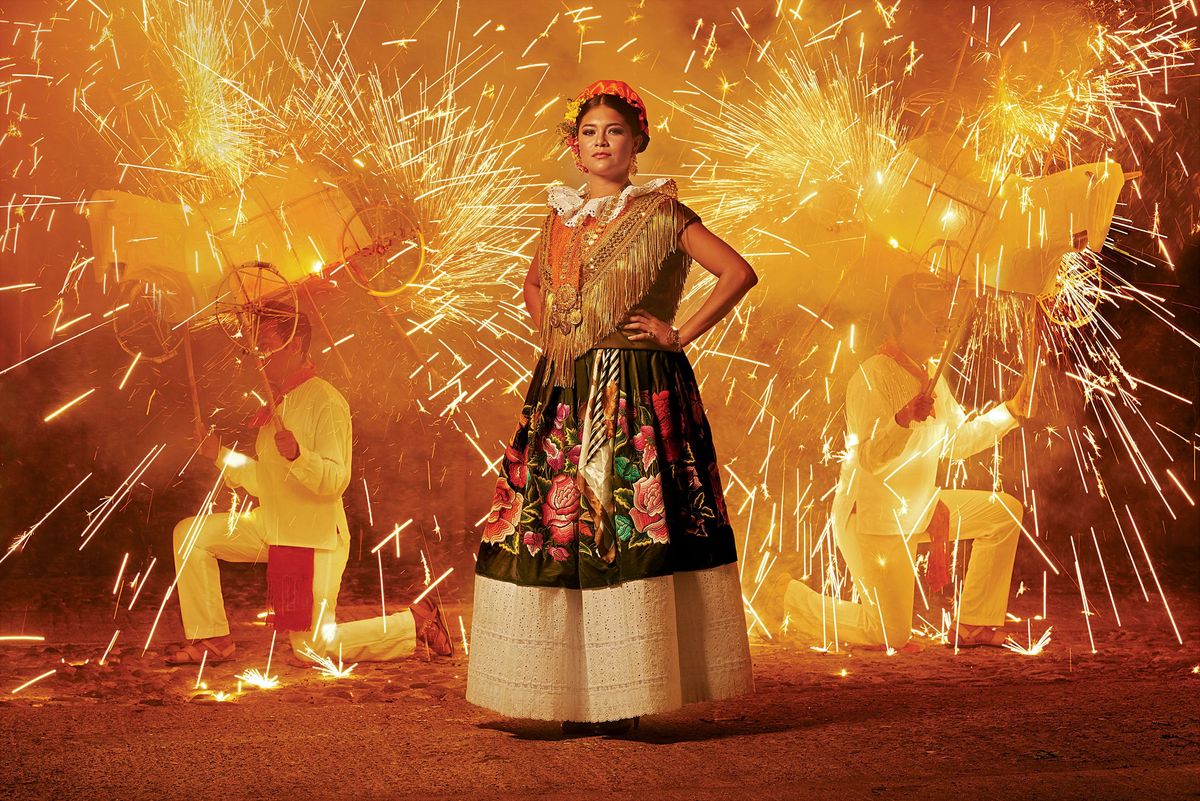 Een vrouw draagt een traditioneel gewaad uit Tehuana De bloemmotieven zijn geborduurd in zijdedraad op fluweel en versierd met goudjuweeltjes die van generatie op generatie worden overgedragen De twee mannen achter de vrouw zijntoritosstiertjes