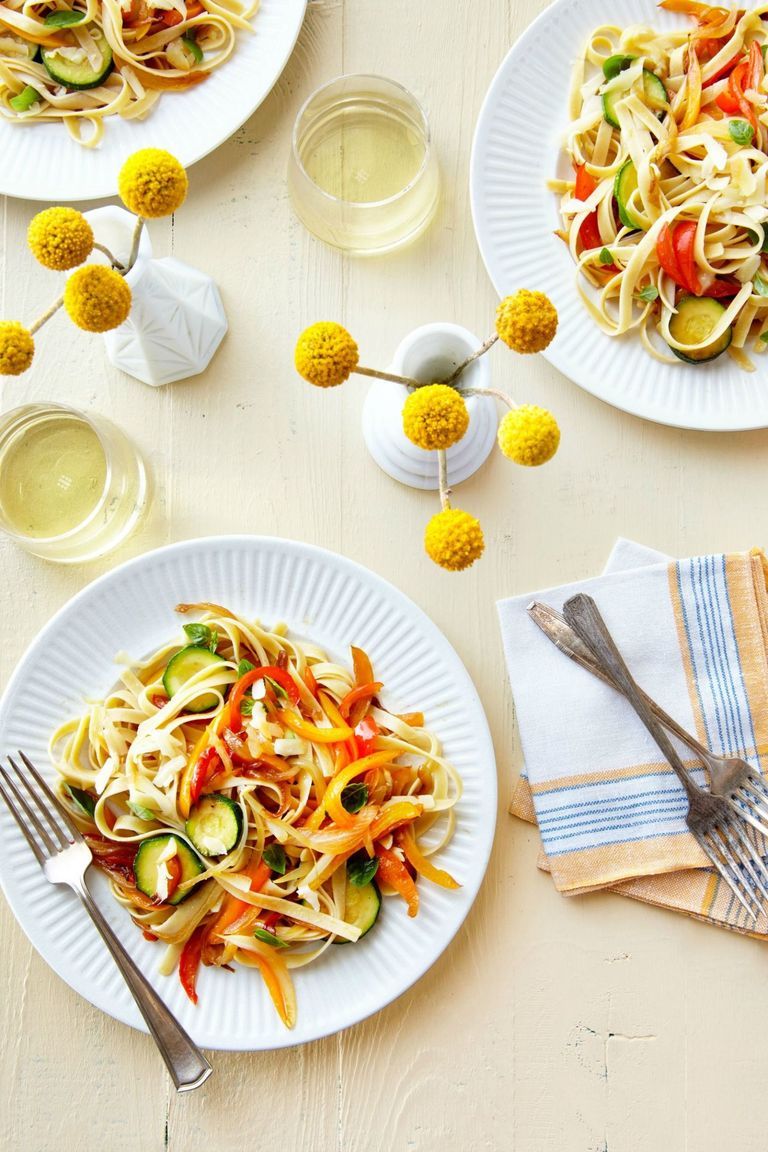 3 plates of fettuccine pasta with colorful vegetables, set on a table with cloth napkins, forks, and milk glass vases of yellow craspedia flowers