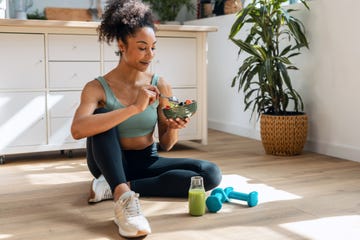 shot of athletic woman eating a healthy fruit bowl while sitting on floor in the kitchen at home